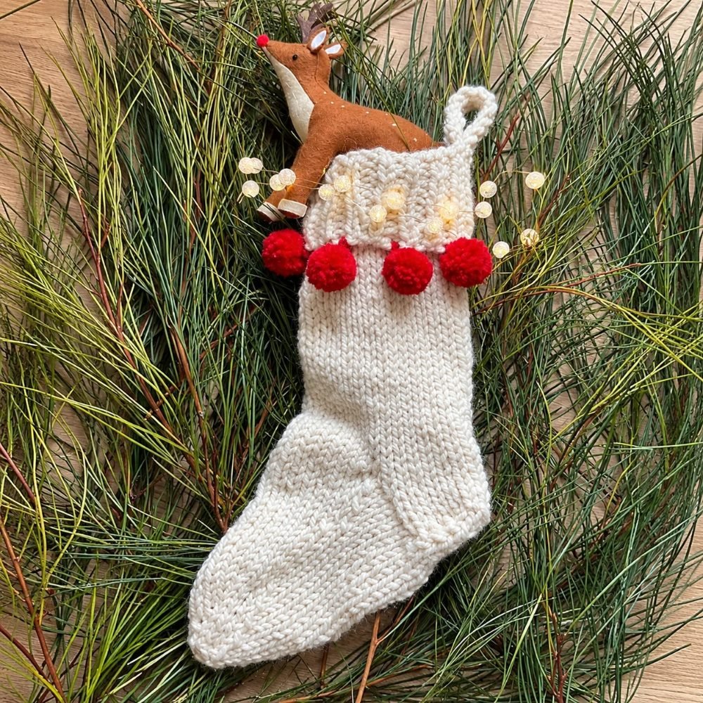 Cream chunky knitted Christmas stocking with red pompoms along the cuff, lying on pine branches, from the How To Knit A Christmas Stocking pattern by Handy Little Me