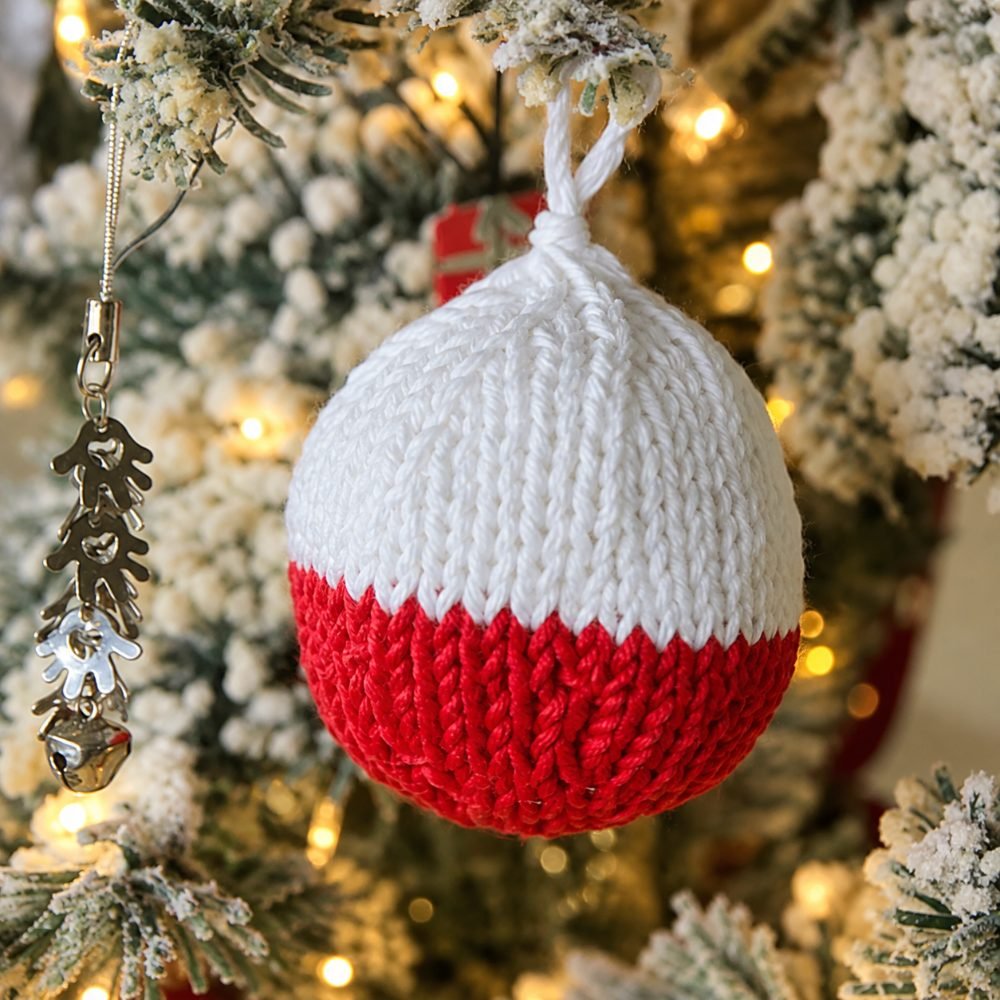 Red and white two-color knitted Christmas ball ornament hanging on a frosted tree, from So Woolly