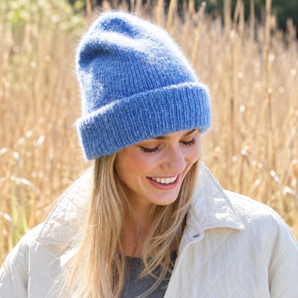 Bright blue knitted beanie with a folded brim worn in a sunny field, DROPS Isle Waters