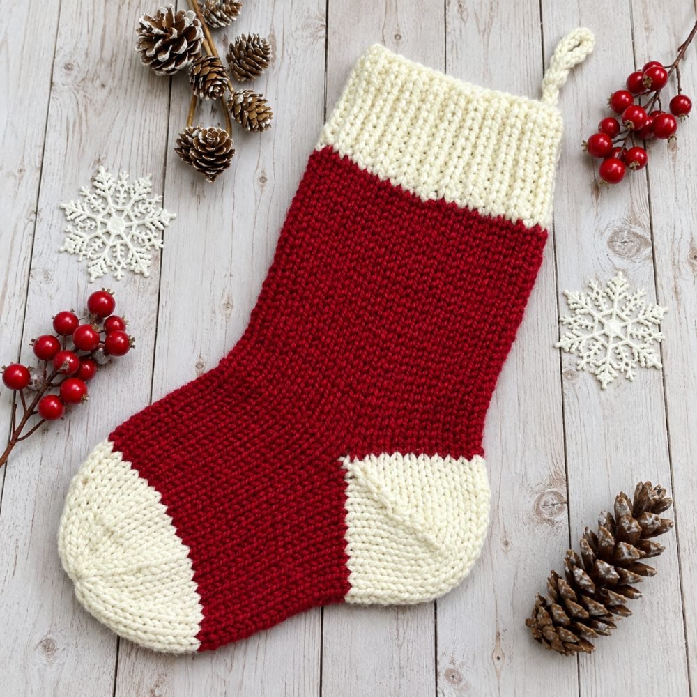 Red knitted Christmas stocking with a cream cuff, heel and toe beside pine cones and berries, from Fox & Pine Stitches