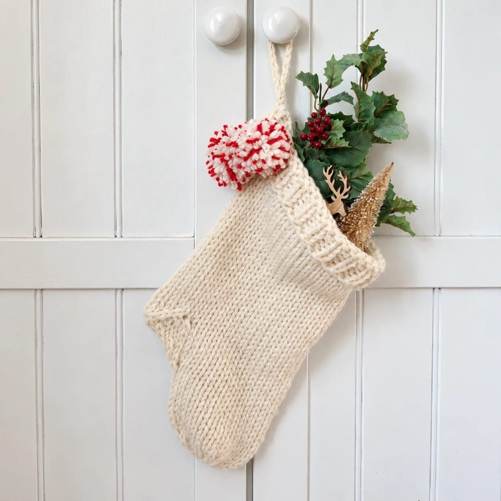 Cream knitted Christmas stocking with pompoms and greenery hanging on a white cupboard door, from A Box of Twine